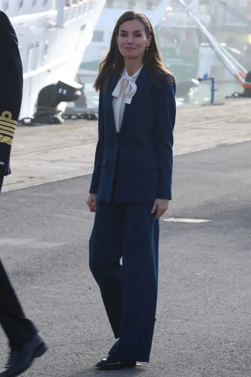 La Reina Letizia apuesta por un traje chaqueta pantalón azul marino y camisa blanca con lazo en el cuello.