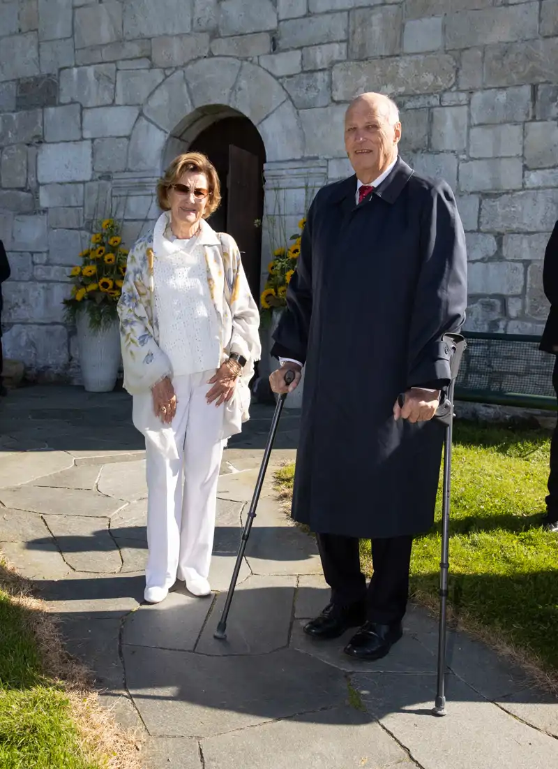 Sonia y Harald de Noruega paseando con girasoles de fondo. El Rey permanece apoyado en unas muletas